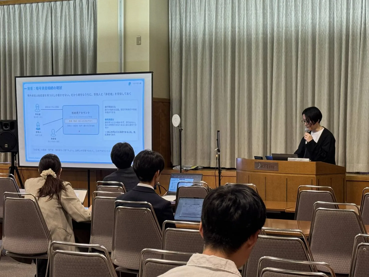 アクトシティ浜松の会場で、「暗号資産相続の現状」のスライドを映しながら当社社員の室橋太郎が発表し、聴講者がノートPCを開いて聞いている様子