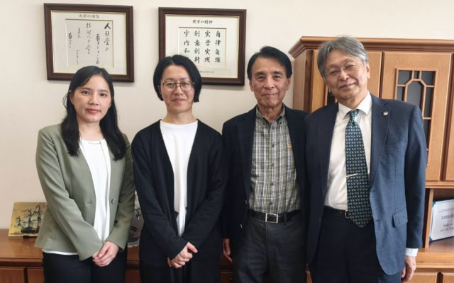 オフィス内で額装された書と棚を背景に、4名が並んで撮影された集合写真